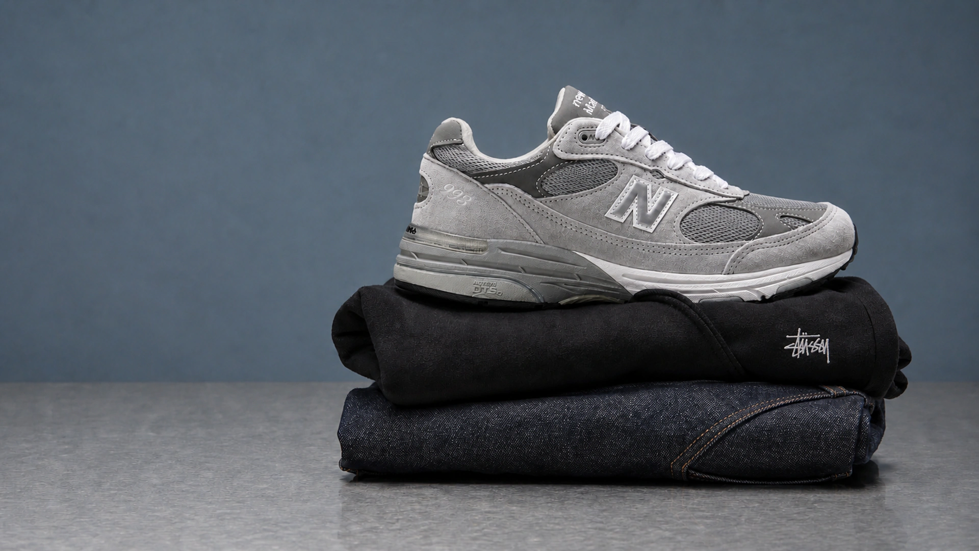 Серые кроссовки New Balance 993, чёрное худи Stüssy и джинсы A.P.C. на сером столе